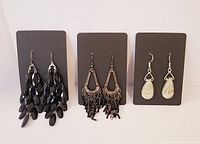 Three pairs of dangle earrings displayed on dark holders, showing black faceted teardrop beads, metal frame and tassels, and light beige stone teardrop shapes.