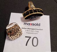 Two statement cocktail rings; left one is silver with circular raised patterns, right one is gold-tone with large black faceted stone and black geometric stones on band, displayed on MaxSold paper.