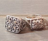 Two square cocktail rings displayed side by side on wood surface; left ring has floral design with gold accents, right features woven pattern