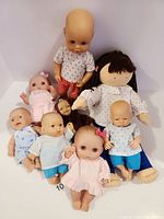 Photo showing seven vintage dolls arranged sitting and standing against a white background with visible details of clothing and facial features.