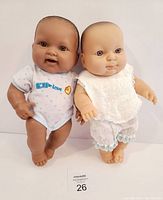 Two baby dolls, one dark-skinned in blue onesie and one light-skinned in white bib and pants, shown standing.