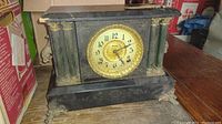 Front side view of the antique mantel clock showing its black body, gold decorative columns, ornate feet, and clock face with Arabic numerals