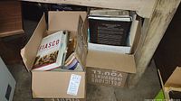 Two cardboard boxes filled with various hardcover and softcover books placed under a table.