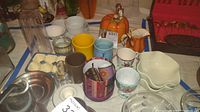 Wide view showing multiple ceramic mugs, egg tray and cups, glass jar, and small bowls grouped together on a table.