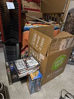 Photo shows two black plastic organizing racks stacked vertically next to a cardboard box labeled Hello Fresh. Several DVDs and VHS tapes are stacked on the carpet floor beside the box and racks.
