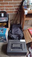 Photo of fishing gear including two fishing rods leaning against a wall, a black carry bag likely containing waders, a black plastic tackle box, and other assorted boxes around.