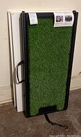 Folding dog ramp covered with artificial turf leaning against wall with two white wooden shelves behind.