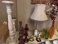 Photo of table lamp with beige shade, various candlesticks, bird figurines, glassware, and tall orange vase on table.