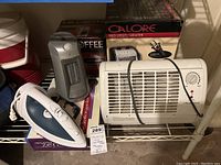 White rectangular electric heater with thermostat control, gray portable heater, blue and white iron, Aroma Zen Garden box and Mr Coffee pot box on metal shelf
