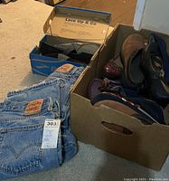 Two pairs of Levi's blue denim jeans with red tags stacked on the floor next to a shoe box labeled 'Lace Up & Go' containing a pair of Vans shoes and a box of five women's shoes in various styles and colors.