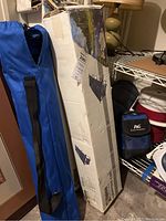 Photo of long blue carry bag with black handles likely containing a camping chair and two white cardboard boxes containing camping cots with black printed graphics.