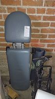 Photo showing the collapsible weight bench with padded head and backrest against a brick wall, next to rolled yoga mat and luggage cart.