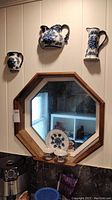 Three half pieces of a blue floral china teapot, coffee pot and pitcher mounted on the wall with a wooden framed octagonal mirror below them, showing their size and arrangement.