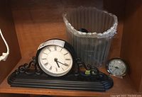 Black composite and metal mantel clock with a round face featuring Roman numerals alongside a small silver-tone Westclock alarm clock and a clear plastic container.