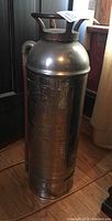 Metal vintage fire extinguisher with tarnish, embossed Arrow branding visible, hose coil by the side.