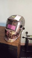 Side view of welding helmet showing camouflage pattern and viewing window with purple-tinted lens.