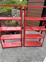 Two bright red metal shelving units shown outside against a natural background. Each has four shelves and cross braces on the back.