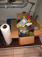 Box with assorted cleaning products including spray bottles and a bleach cleanser can, beside a roll of paper towels on a stand.