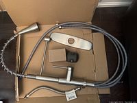 One handle brushed nickel kitchen faucet with flexible pull-down sprayer, mounting plate, weight, and braided supply line laid out on cardboard base.