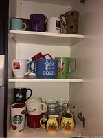 Top shelf of cabinet showing nine assorted mugs with various colors and designs, including purple, white, brown with dots, blue with text, green with cartoon, and branded mugs.