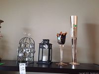 Four decorative items displayed on a shelf: two black metal candle holders with circular details and glass cups, a black square lantern candle holder, a Murano glass candle holder with twisted clear glass stem and dark top with gold accents, and a tall slim silver metal vase with etched designs.