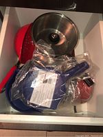 Top view of various pots and pans including a metal pot with lid, blue Small Micro Cooker in packaging, and red handled cookware.