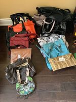 Photo shows approximately thirteen assorted purses and bags in various colors and styles placed on a wooden floor.