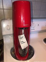 Front view of red Sodastream soda maker with clear bottle, placed on stove with maxsold tag attached.