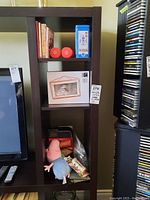 Photo showing section of shelving with various items including boxed Umbra photo display, blue tin box, books, Scrabble game, plush toy, and other small items.