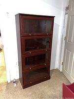 Front view of the wood barrister bookcase showing four glass-front compartments with brass knobs, placed against a white wall near a door.