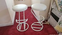 Two vintage white vinyl padded round seat bar stools with white enameled metal frames, photographed together on a red carpet.