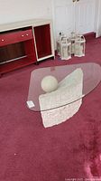 Pedestal coffee table on maroon carpet with white textured stone base and ball, clear thick glass top with rounded corners visible from front angle.
