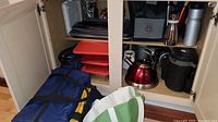 Open cabinet showing red stainless steel tea kettle, black insulated travel mugs and bottles, red and black cookie sheets, and insulated lunch bags in blue and green on floor and shelf.