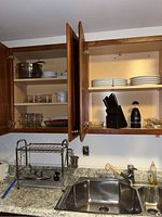 Kitchen cabinet open showing stacked white porcelain plates, glass mugs, CorningWare mugs, black knife block with cutlery and Power Chopper XL. Metal wire drying rack is on countertop by sink.