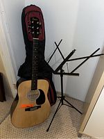 Full view of the acoustic guitar with its soft case leaning against the wall next to a black metal sheet music stand.