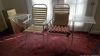 Two white metal lawn chairs between two folding tray tables and a gold-tone stand in corner