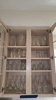 Glass cabinet shelves with multiple Mikasa glassware items including wine glasses, rocks glasses, glass mugs, decanter, ice bucket, and serving bowl.