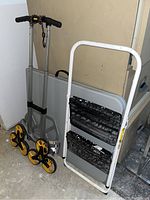 Hand truck with three yellow and black stair-climbing wheels per side, white folding step stool, and folded gray catering table.