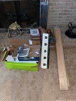 Full lot overview showing boxes with hardware, wall rack leaning, wooden boards, and thermostat box on carpeted floor near brick fireplace.