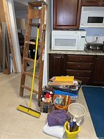 Photo showing a 6-foot wooden ladder leaning against a wall next to a kitchen counter, yellow push broom, blue box with painting brushes, rollers, paint tape, two buckets, orange container and cloths.
