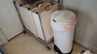 Rolling 3-bag laundry sorter with beige fabric bags on metal frame and white foot pedal trash bin side by side.