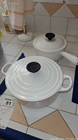 Two white Le Creuset pots with lids placed on a tiled kitchen countertop, showing black knobs on lids and overall condition.