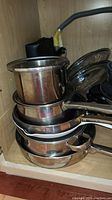 Stack of stainless steel pots and pans in a cabinet, showing lids and cookware handles.