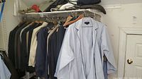 Men's suits and blazers hanging on a metal rod inside a closet space. Variety of colors including black, navy, and beige.
