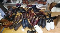 Wide view showing a variety of men's shoes including boots, dress shoes, sneakers, and slides arranged on floor and shelves.