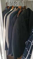 Photo showing several men's suit jackets hanging on a rod, including dark, navy, grey and houndstooth pattern, various styles hung on wooden and plastic hangers.