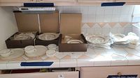Full lot showing multiple pieces of Mikasa Antique Lace dishware including plates, cups, saucers, and bowls wrapped in protective packaging and placed on kitchen counter.
