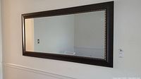 Full view of large rectangular mirror mounted on wall, showing reflective surface and dark brown frame with gold beaded trim.