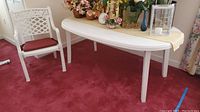 White plastic semi-oval patio table with decorative items on top and a white patio chair with red cushion beside it on red carpet.