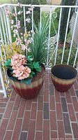 Two large glazed ceramic planters on a porch, one with an arrangement of faux flowers including pink blossoms, hydrangea, grass-like green foliage and yellow leaves, the other empty.
